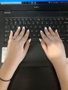 Τα χέρια της Χριστίνας ακουμπισμένα επάνω στο πληκτρολόγιο ενός μαύρου λάπτοπ. Τα νύχια της είναι βαμμένα λευκά, ενώ δύο νύχια σε κάθε χέρι έχουν επίσης και ασημένιο Γκλίτερ.